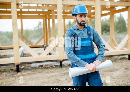 Builder in blue overalls and hard hat with blueprints on the construction site, building wooden frame house Stock Photo