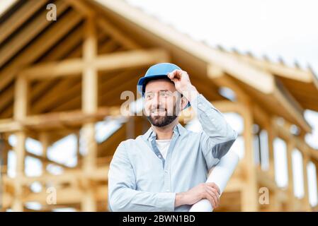 Portrait of an architect or builder in hard hat standing in front of the wooden house structure. Building and designing wooden frame house concept Stock Photo