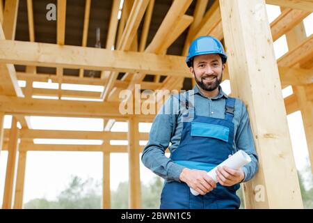 Portrait of a handsome builder in blue overalls and hard hat with blueprints on the construction site, building wooden frame house Stock Photo