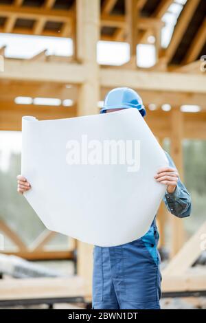 Builder in blue overalls and hard hat looking on the blueprints on the construction site. Building wooden frame house concept Stock Photo