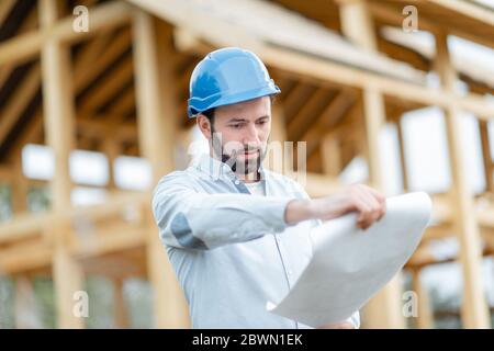 Builder or architect in hard hat supervising a project, standing with blueprints on the construction site. Building wooden frame house concept Stock Photo