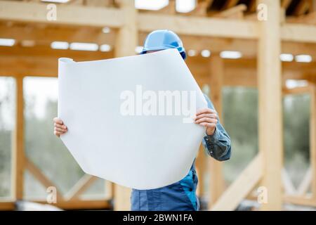Builder in blue overalls and hard hat looking on the blueprints on the construction site. Building wooden frame house concept Stock Photo