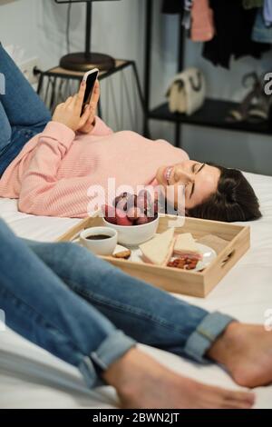 Young couple having breakfast and relaxing while lying on bed in house. Stock Photo