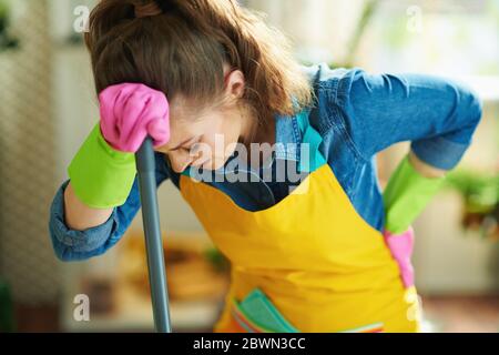 unhappy modern middle age housewife in orange apron and pink rubber gloves with mop having back pain at modern home in sunny day. Stock Photo