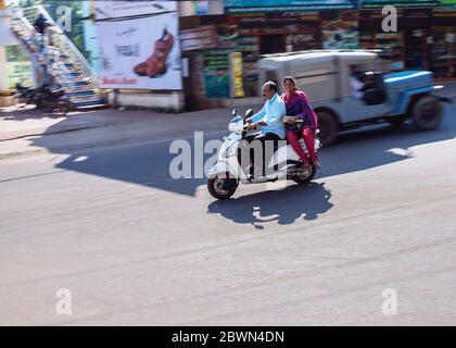 Man keeps eyes on road while speeding by on white scooter with a smiling woman passenger sitting sidesaddle and smiling directly into camera Stock Photo