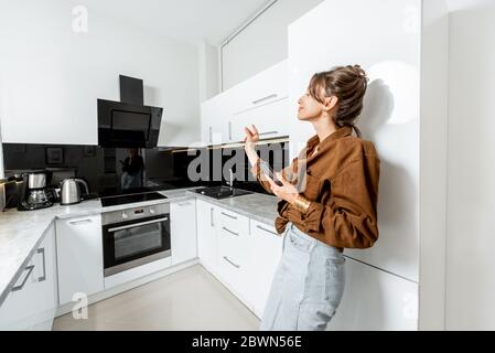 Young woman controlling kitchen appliances with mobile phone and voice commands, wide interior view. Smart home concept Stock Photo