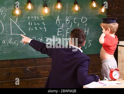 Teacher pointing at the green board while explaining shapes to child. Funny kid in graduation cap pulling up red T-shirt. Man turned back and boy side view in the classroom Stock Photo