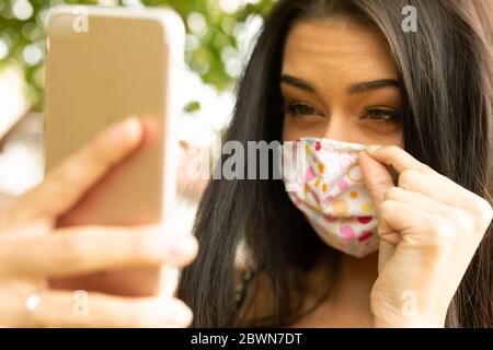 Young woman adjusting the fit of her fashionable textile face mask using her mobile phone worn as protection against the spread of the coronavirus dur Stock Photo