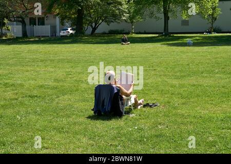 An elderly man sitting alone on the grass in a park while reading a newspaper, Vancouver, BC, Canada Stock Photo