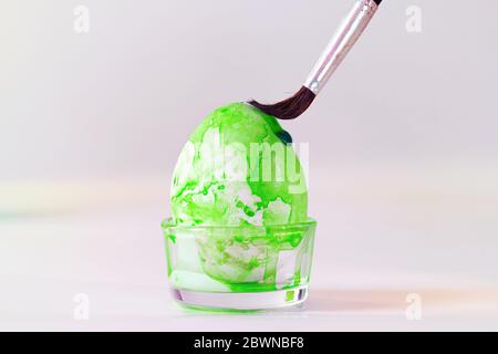 Brush is painting green watercolor on an Easter egg for the holiday decoration, light grey background with copy space, selected focus, narrow depth of Stock Photo