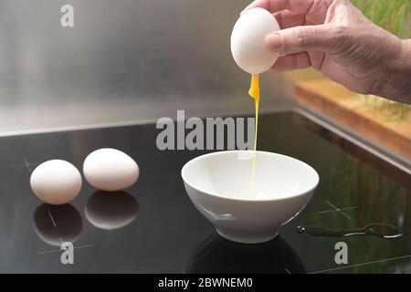 Hand is holding a whole egg shell with leaking yolk over a white bowl, preparation for Easter decoration, copy space, selected focus, narrow depth of Stock Photo