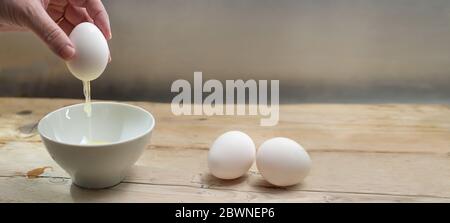 Hand of an older woman is holding a whole egg shell while the protein is running into a white bowl on rustic wood, preparation for Easter decoration, Stock Photo