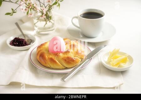 Easter breakfast for one, plaited yeast bread nest with a pink egg, coffee, butter and jam on a white table, during the coronavirus crisis, many have Stock Photo
