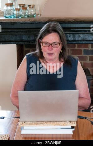 A middle-aged Caucasian woman chatting to friends on a Zoom conference call or cloud meeting from a laptop in her kitchen during the COVID-19 lockdown Stock Photo