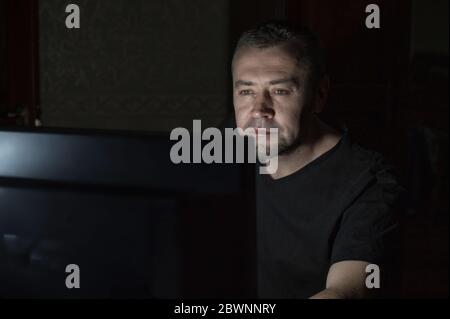 Man works on a computer at night. Attractive man sitting at desk table looking at laptop computer in dark room late at night. Stock Photo