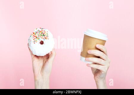 Woman hand holds paper cup with coffee and donut with white icing and multicolored sugar pastry topping, pink background Stock Photo