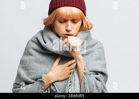 Cute sick young teen woman in red hat, wrapped in warm scarf coughing, closed eyes.Female feeling the first symptoms of illness.Bronchitis, upper resp Stock Photo