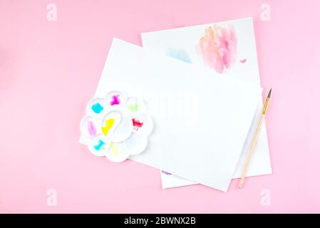 Notebook mock up for artwork with watercolor paints, palette and a paintbrushes. View from above. Artistic work tools on pink table. Stock Photo