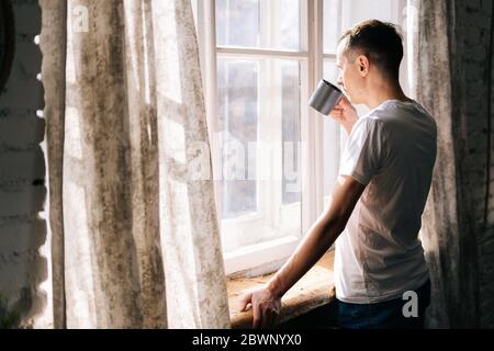 Pensive young man stands by the window on sunny day and takes sip of coffee. Stock Photo