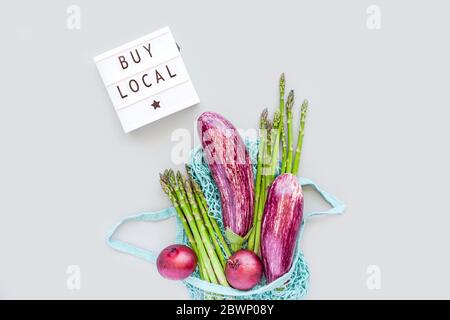 Fresh organic vegetables in eco cotton mesh shopping bag with text Buy Local on lightbox flat lay, top view with copy space on gray background. Sustai Stock Photo