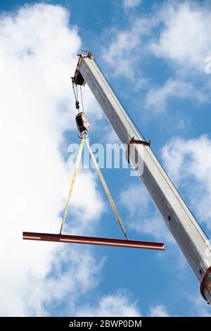 Construction crane with a hook and a metal profile hanging on ropes lifting up, low angle view with cloudy sunny blue sky Stock Photo