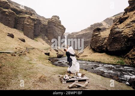 Destination Iceland wedding, near Kvernufoss waterfall. Wedding couple on the shore of a mountain river. The groom wears the bride in a woolen blanket Stock Photo