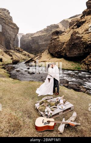 Destination Iceland wedding, near Kvernufoss waterfall. A wedding couple stands under a plaid near a mountain river. The groom hugs bride. They built Stock Photo