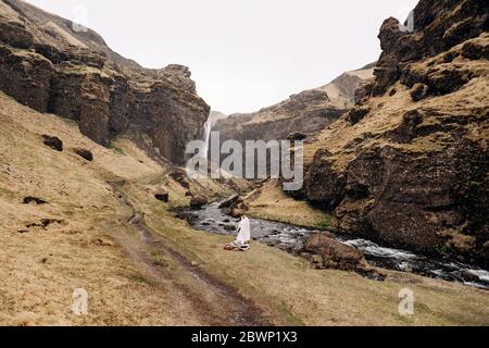 Destination Iceland wedding, near Kvernufoss waterfall. A wedding couple stands under a plaid near a mountain river. The groom hugs bride. They built Stock Photo