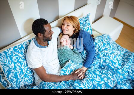 Happy multi ethnic family hugs in bed as soon as they wake up, Caucasian mom and African dad hug their daughter tenderly, cuddles in bed in the mornin Stock Photo