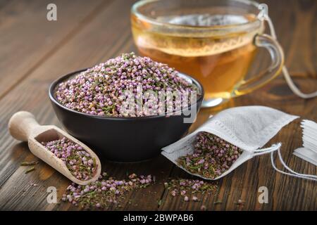 Black ceramic bowl of dry healthy heather, tea bag with Еrica flowers. Glass tea cup with herbal teabag inside on table. Alternative medicine. Stock Photo