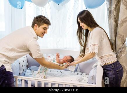 Young couple putting crying newborn son to sleep in white crib next to bunch of balloons Stock Photo