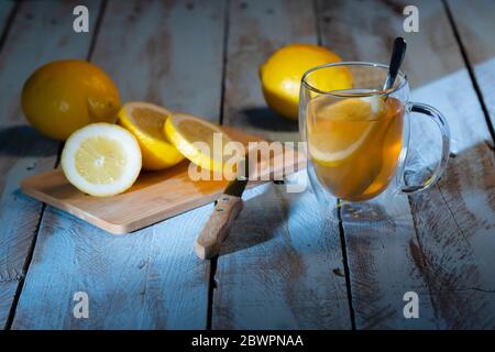 Delicious cup of tea with a lemon slice next to a cutting board with a sliced lemon and a knife, with some whole lemons at the back on a wooden table. Stock Photo