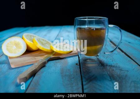 Close up of a cup of tea next to a cutting board with a sliced lemon and a knife on a wooden board on an out of focus background. Relaxation and lifes Stock Photo