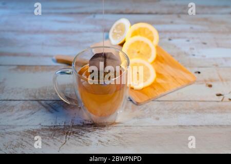 Close up of a cup of tea with a dipping tea bag and an out of focus cutting board with a sliced lemon at the back on a wooden table. Relaxation and li Stock Photo
