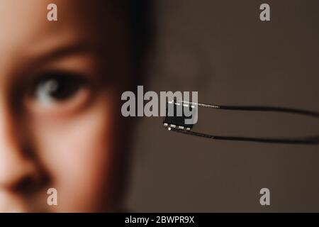 A pair of tweezers holds a black identification microchip against the background of the face. The concept of chipping people after the coronavirus epidemic. Stock Photo