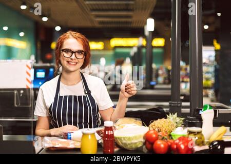 Young smiling female cashier in eyeglasses and striped apron with products on cash line happily looking in camera showing thumb up gesture while Stock Photo