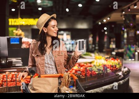 Nice smiling girl in hat with trolley full of products happily showing new cellphone on camera in supermarket Stock Photo