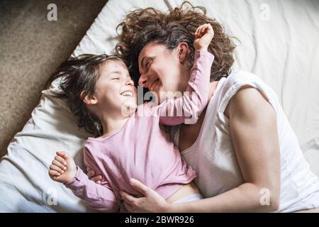 A mother and her daughter smile as they caress each other and play around on a bed.