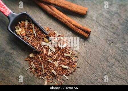 Japanese measuring tea spoon spreads aromatic rooibos dried leaves on wooden surface. Cinnamon bark strips on the side Stock Photo