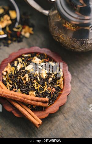 Aromatic fruity black tea dry leaves on small wooden tray, metal kettle and cinnamon bark strips on a side set on a wooden surface Stock Photo