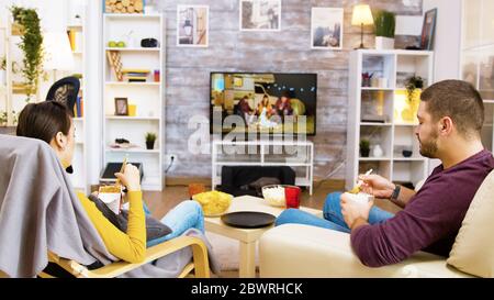 Back view of young couple sitting on comfortable chairs eating noodles in front of tv using chopsticks. Cat sleeping on her cozy bed. Stock Photo