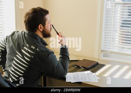 Thinking bearded businessman sitting at office with documents, tablet on table. Stock Photo
