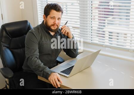 Thinking employee sitting with pen and laptop at cabinet. Stock Photo
