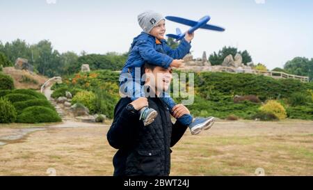 Portrait of smiling toddler boy sitting on father's shoulder at park and launching toy airplane Stock Photo