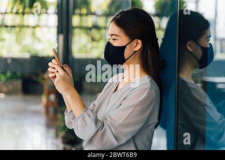 Young beautiful Asian woman with surgical mask for face protection using a smartphone at office building Stock Photo
