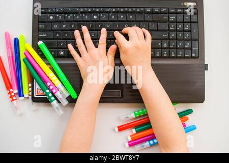Child's typing on a laptop keyboard with colorful pencils and markers on a whiet table. Flat lay style. Homeschooling concept. Stock Photo