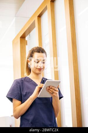 Low angle of young woman in scrubs smiling and browsing data on smartphone while working in contemporary clinic Stock Photo