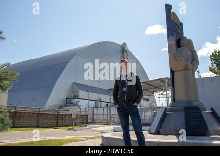 Young adult man posing in front of the chernobyl sarcophagus structure, huge metal confinment unit. Standing next to the monument Stock Photo