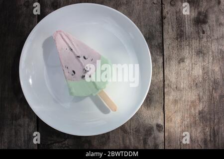 Pink ice cream popsicle in the shape of watermelon on a white plate. Vintage wooden table background with copy space. Summer sweets and desserts. Stock Photo