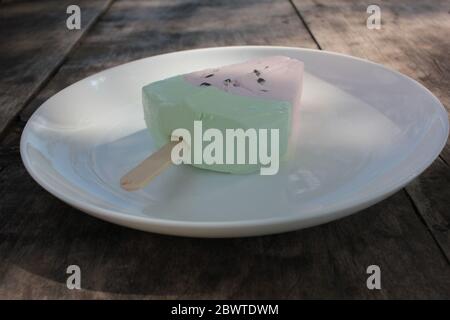 Pink ice cream popsicle in the shape of watermelon on a white plate. Vintage wooden table background with copy space. Summer sweets and desserts. Stock Photo